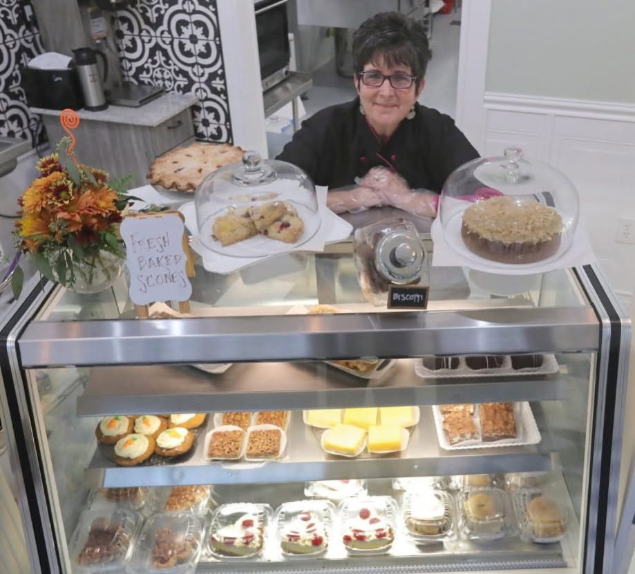 Oh So Sweet bakery owner Judy Pierce poses behind a pastry case filled with desserts and pastries. 